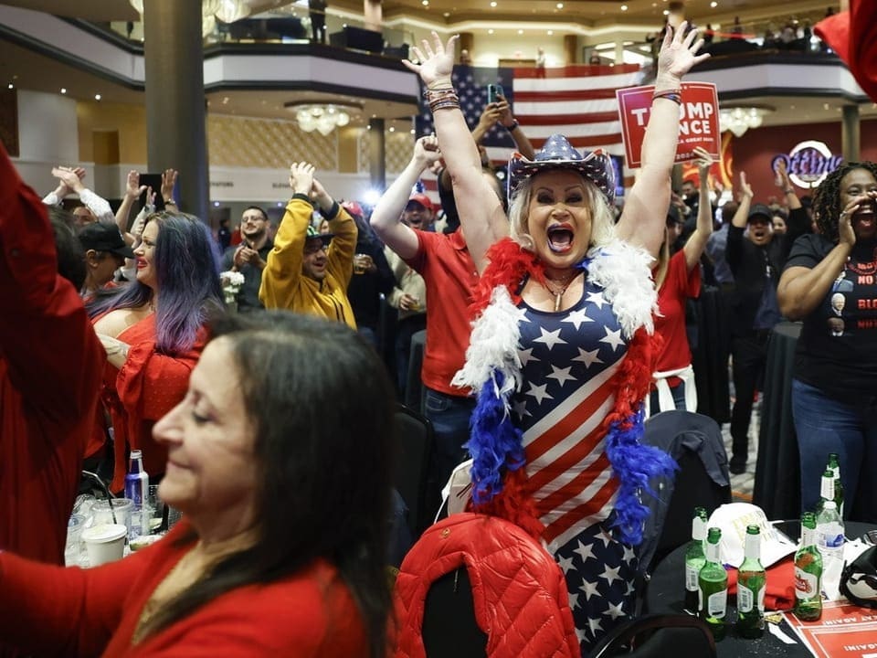 Menschen jubeln bei einer Veranstaltung, Frau in USA-Flaggenkleidung im Vordergrund.