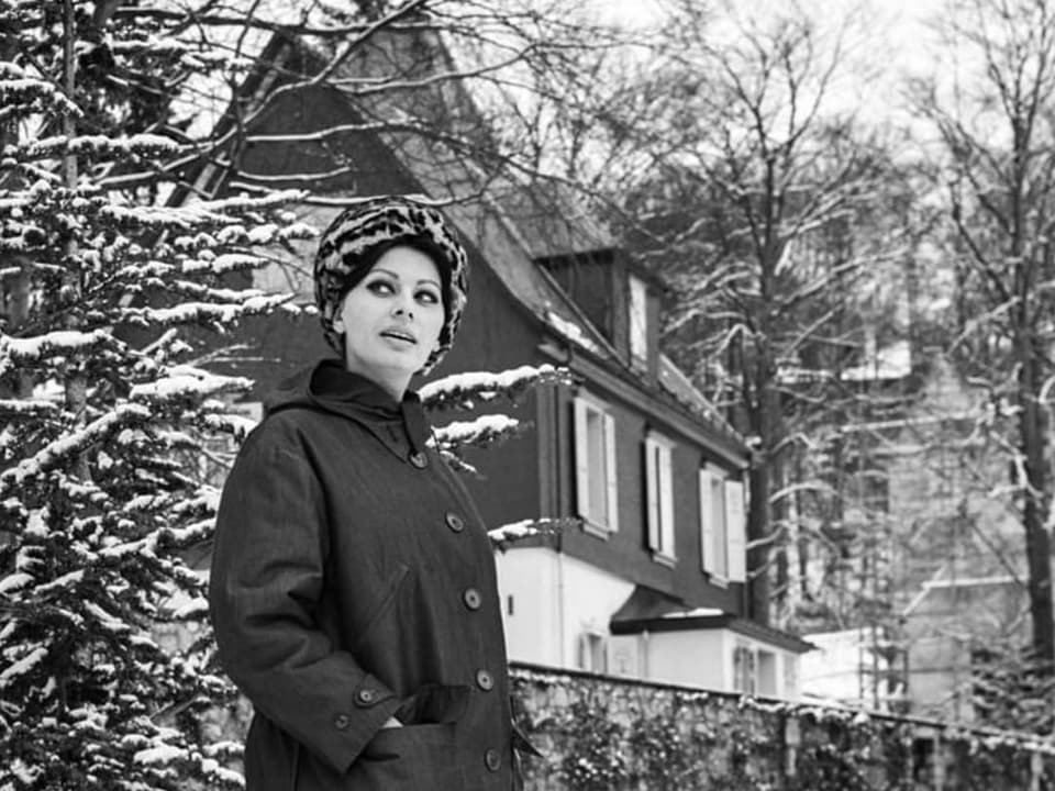 Eine Frau mit dunkel geschminkten Augen steht im Schnee vor einem grossen Haus.