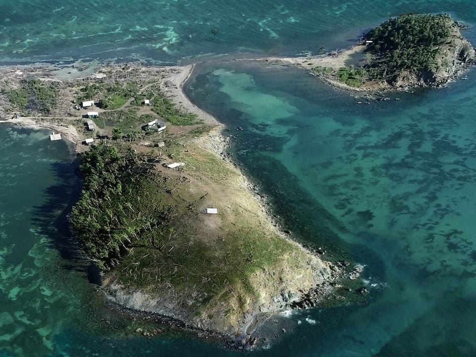 Luftbild einer kleinen, bewaldeten Insel mit Hütten in türkisfarbenem Wasser.