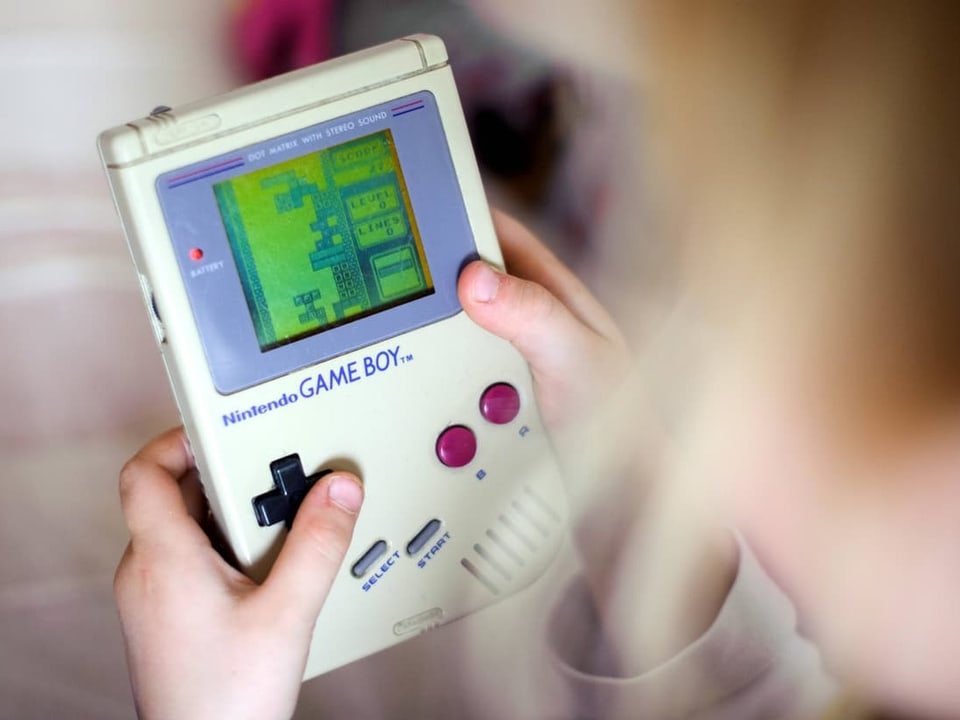 Ein Kind hält einen grauen GameBoy, darauf läuft das Spiel Tetris. Die Blöcke sind in Grautönen.