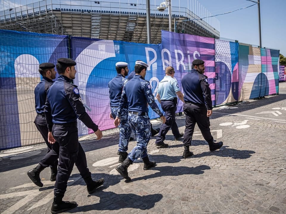 Sechs uniformierte Männer gehen vor einer Paris 2024-Werbung.