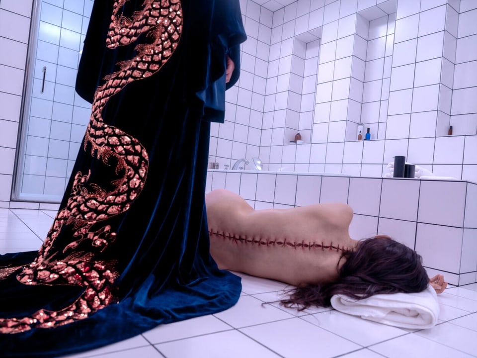 A woman lies naked with a stitched back on the floor of a white-tiled bathroom.