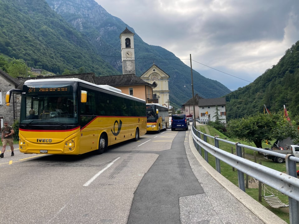 Gelber Bus auf einer Strasse vor einer Kirche in einer bergigen Landschaft.