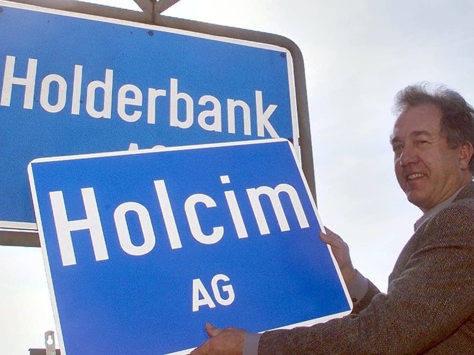 Mann zeigt auf Schild mit Aufschrift 'Holcim AG' vor Schild mit Aufschrift 'Holderbank'.