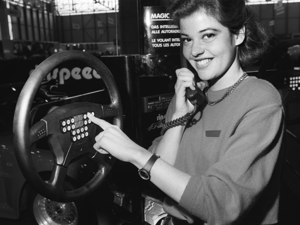 Frau lächelt und zeigt auf Lenkradmodell mit eingebautem Telefon in einer Ausstellung.
