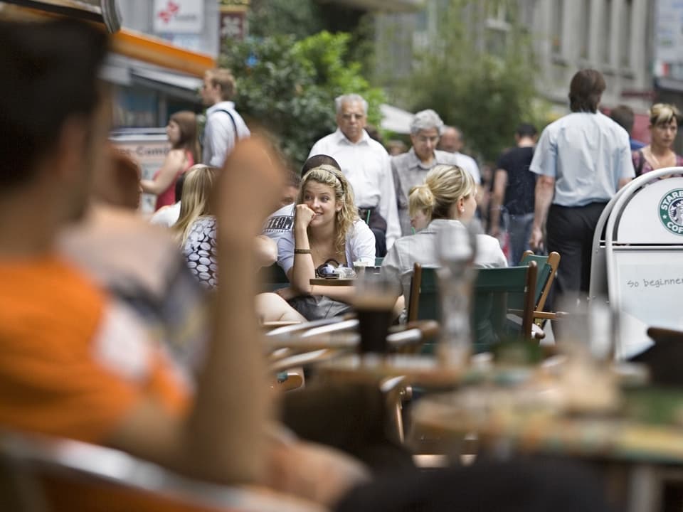 Leuten sitzen an Restauranttischen, andere spazieren vorbei.