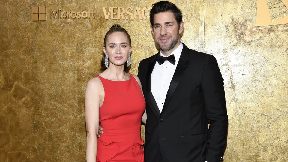 Emily Blunt im roten Kleid und John Krasinski mit schwarzem Sacko und Fliege posieren vor einer Fotowand.