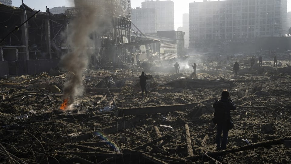 Foto von einer zerstörten Fläche, überall liegen braun-graue Trümmer, Rauch steigt auf. Menschen auf dem Trümmerfeld.