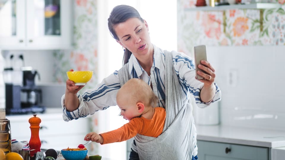 Auf dem Bild ist eine Frau mit einem Baby in der Tragetasche zu sehen. Gleichzeitig hält sie ein Handy in der Hand.