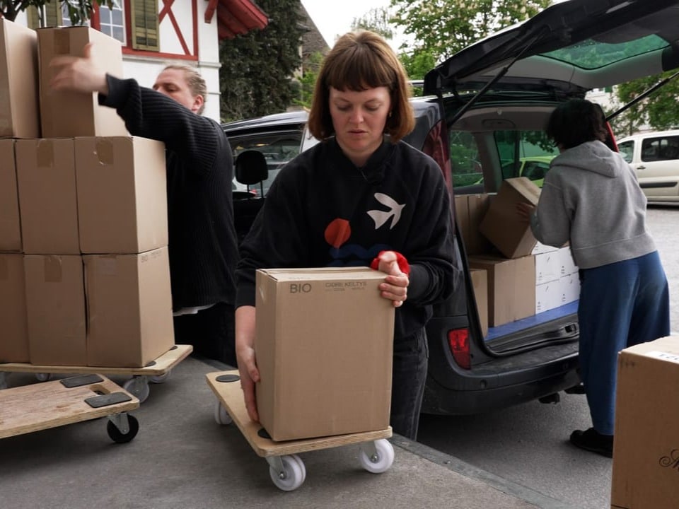 Drei junge Menschen transportieren Weinkisten vom Lager in den Kofferraum eines Autos.