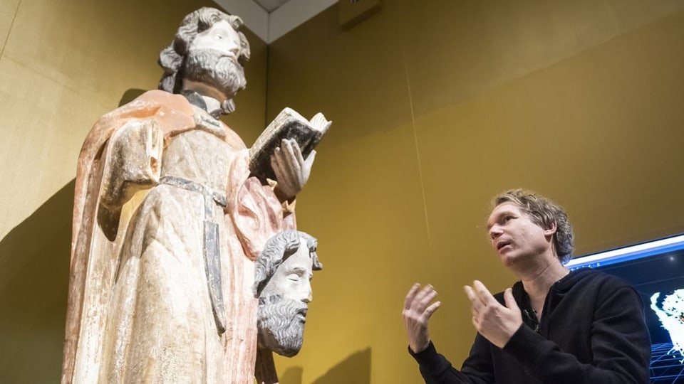 Links sieht man eine Holzfigur, überlebensgross; rechts daneben steht ein Mann und gestikuliert.