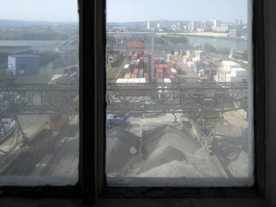 Durch ein staubiges Fenster sieht man im Hintergrund den Rhein, davor Containertürme und graue Berge an Rohstoffen.