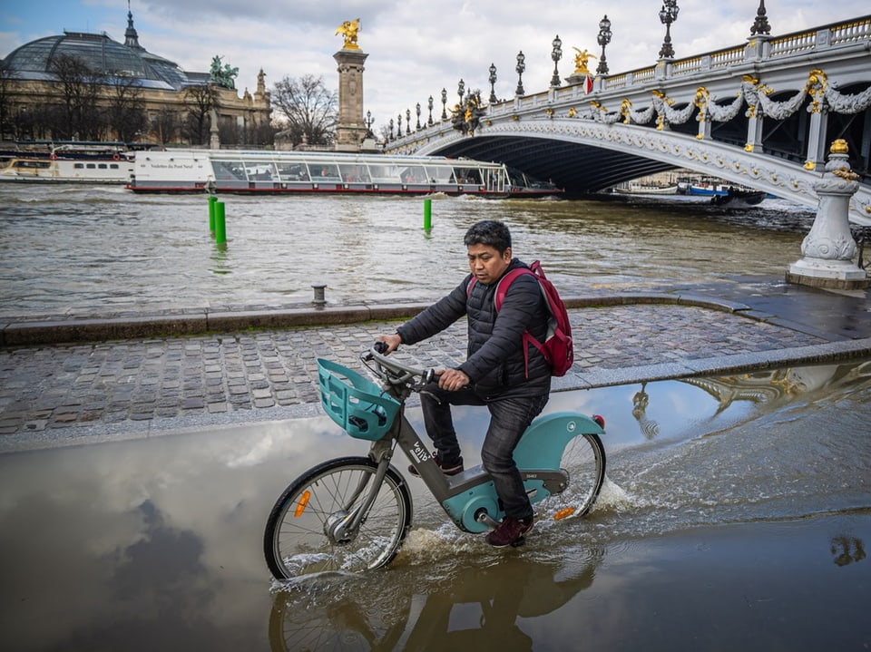 Mann fährt Fahrrad durch überfluteten Bereich an der Seine bei Brücke.