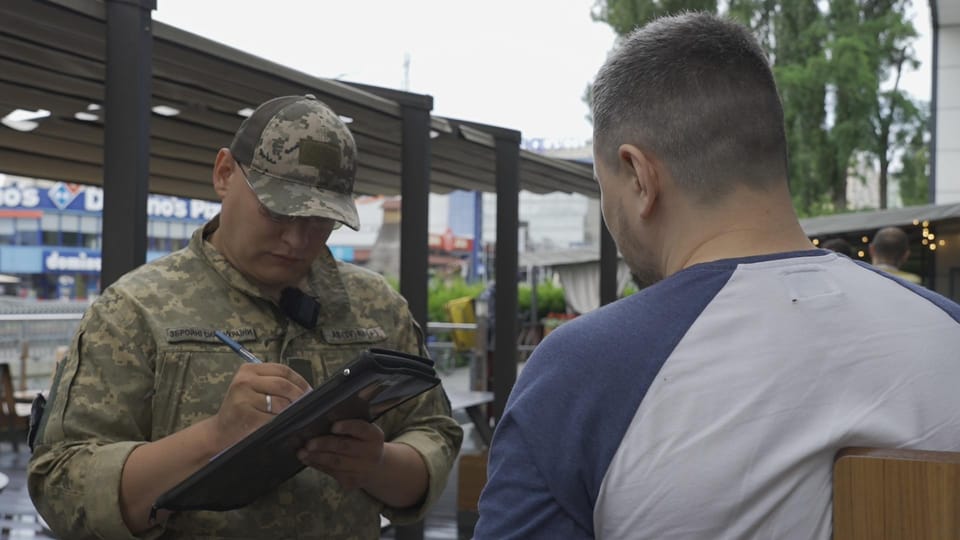 Soldat in Tarnkleidung schreibt auf ein Klemmbrett vor einem sitzenden Mann.