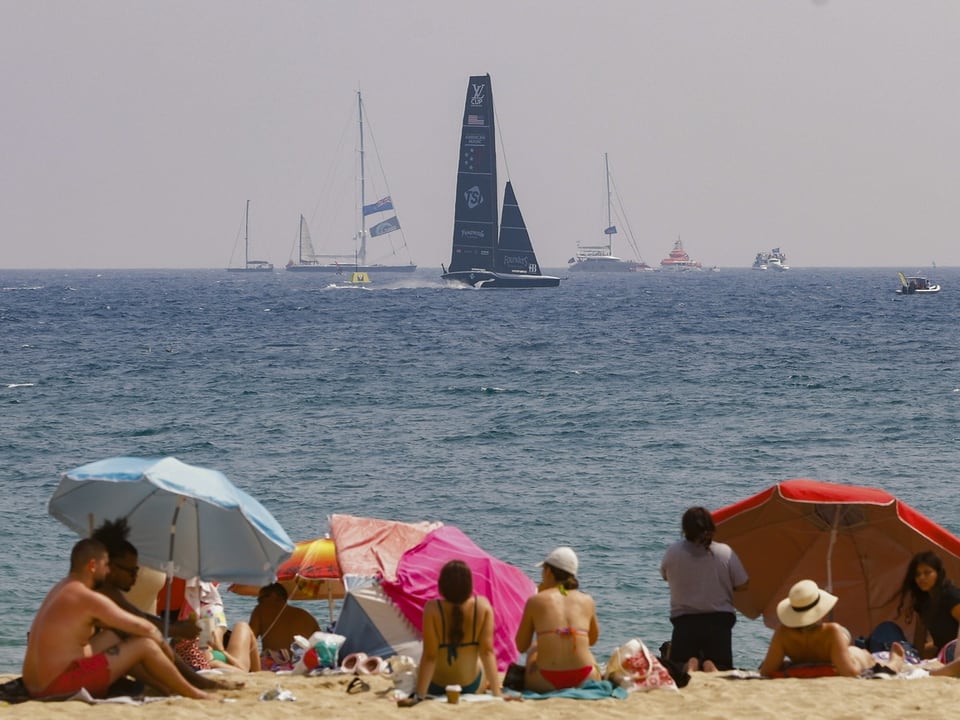 Menschen am Strand mit Sonnenschirmen und Segelbooten im Meer.