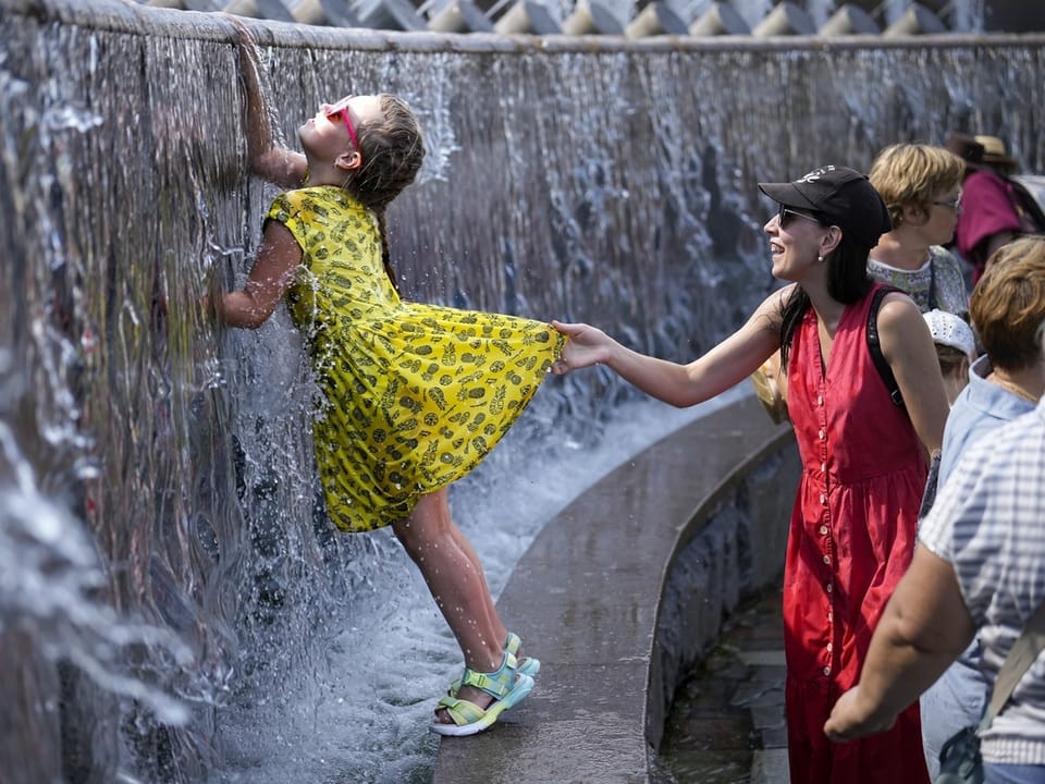 Kind in gelbem Kleid spielt in einem Wasserfall, Frau hält das Kleid fest.