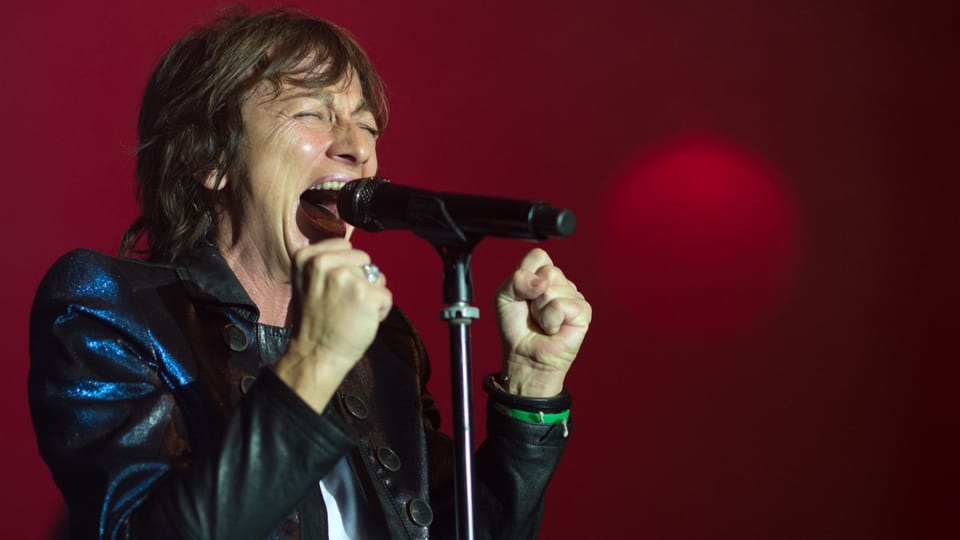 A woman wearing a leather jacket sings into a microphone in front of a red wall. 