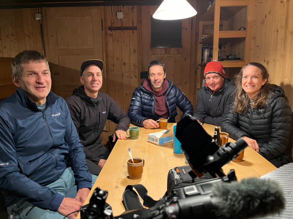 Fünf Personen sitzen lächelnd an einem Holztisch in einer gemütlichen Hütte.