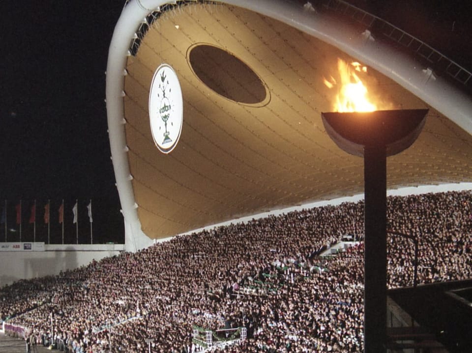 Grosses Stadion bei Nacht mit brennender olympischer Flamme und Menschenmenge.