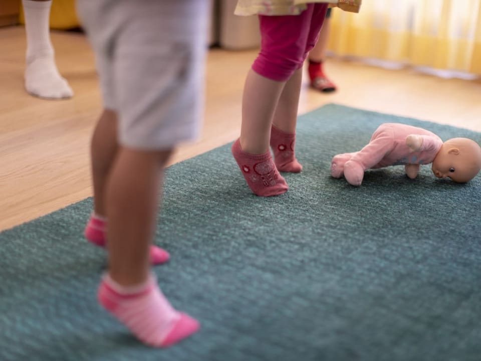 Bild von Kinderbeinen in einer Kindertagesstätte. Auf dem Teppich liegt eine Baby-Puppe.