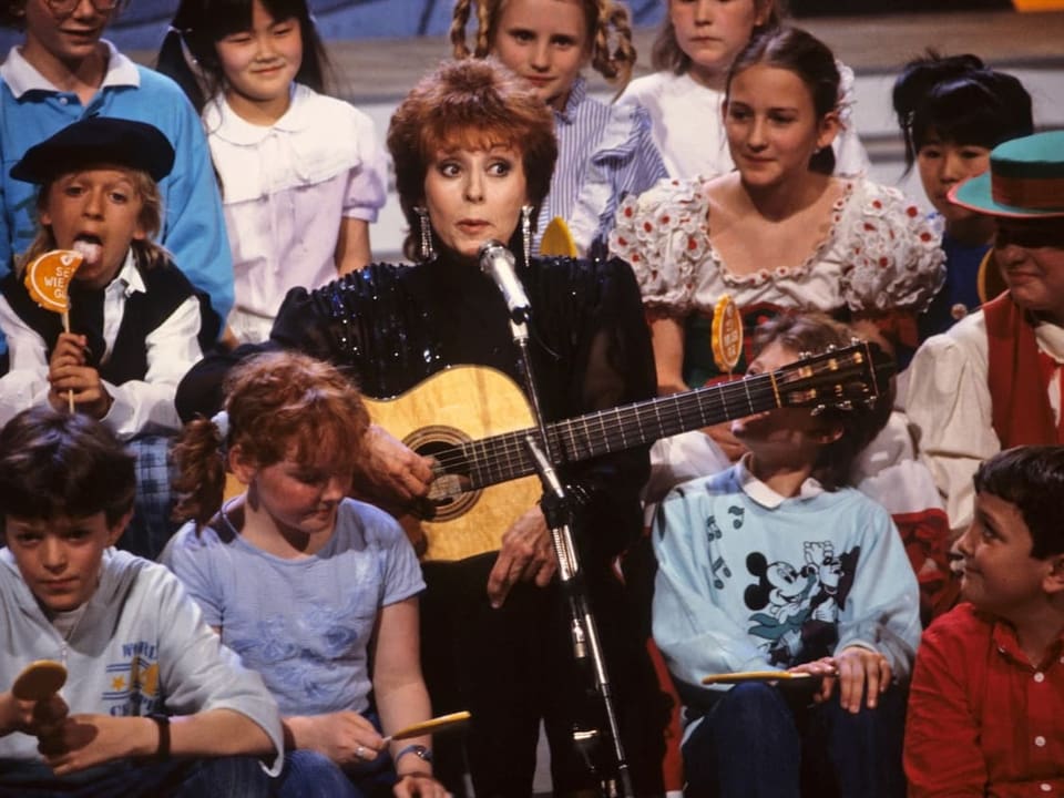 Frau mit Gitarre, umgeben von Kindern auf einer Bühne.