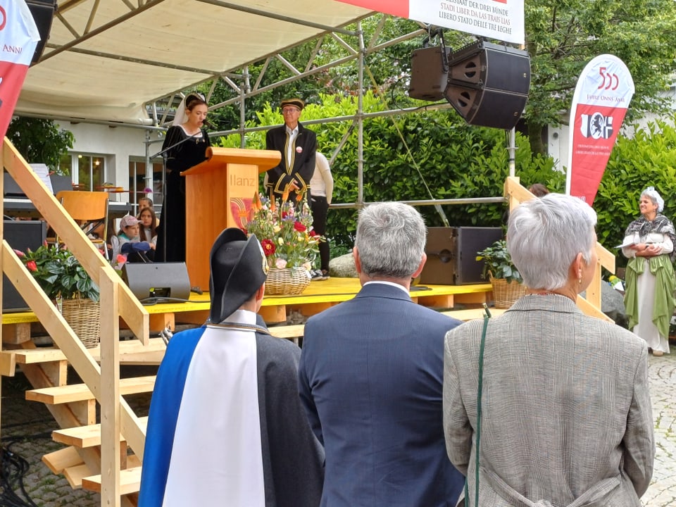 Menschen beobachten eine Rednerin und einen Mann auf einer Bühne im Freien.