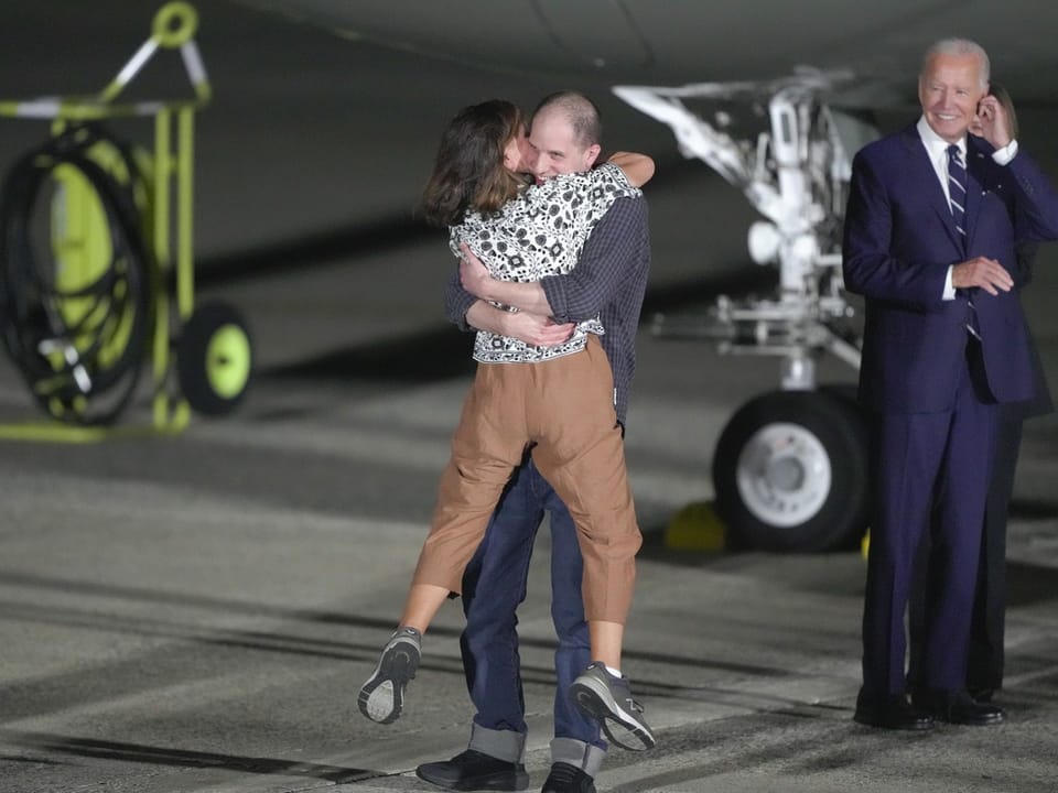 Eine Frau und ein Mann umarmen sich, während ein älterer Mann in einem Anzug in der Nähe eines Flugzeugs steht.