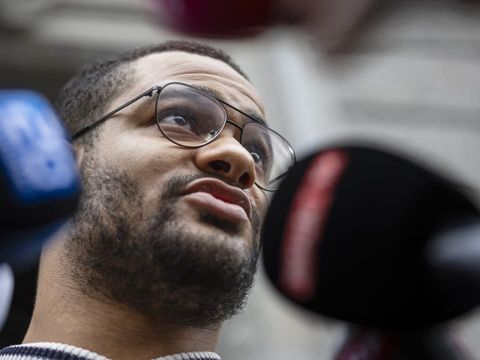 Ein junger Mann mit Brille spricht in zwei Pressemikrofone