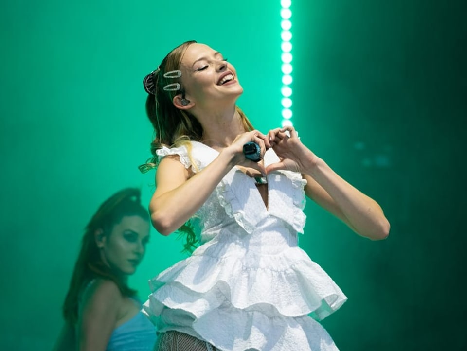Sängerin Angèle steht auf der Bühne und formt ein Herz mit ihren Fingern in Richtung Publikum