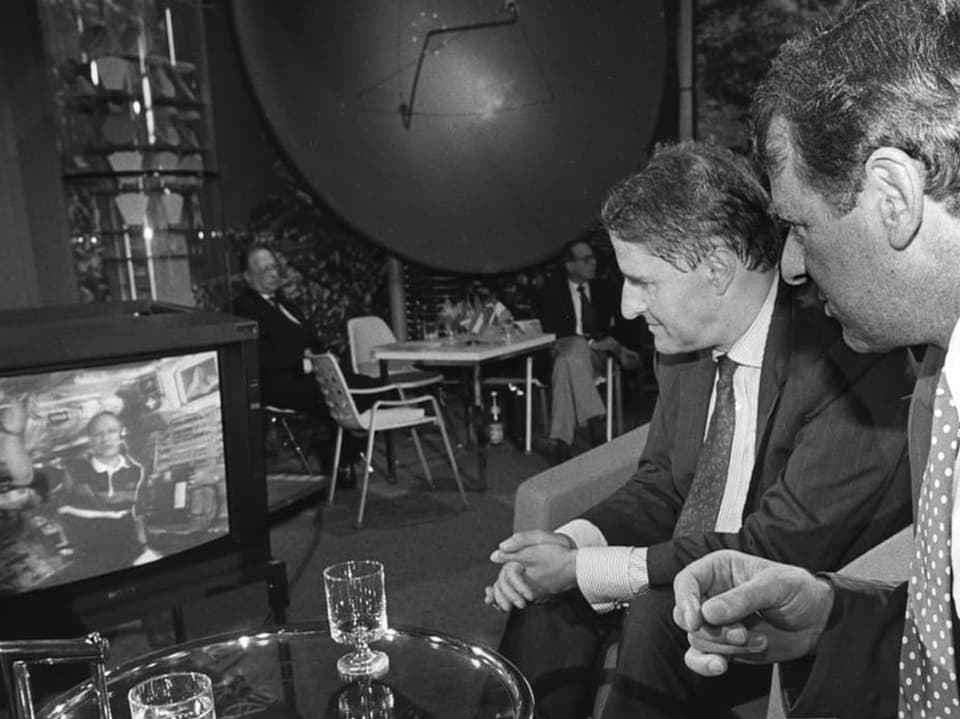 Zwei Männer sehen auf einen Fernseher, der eine Person im Raumanzug zeigt.