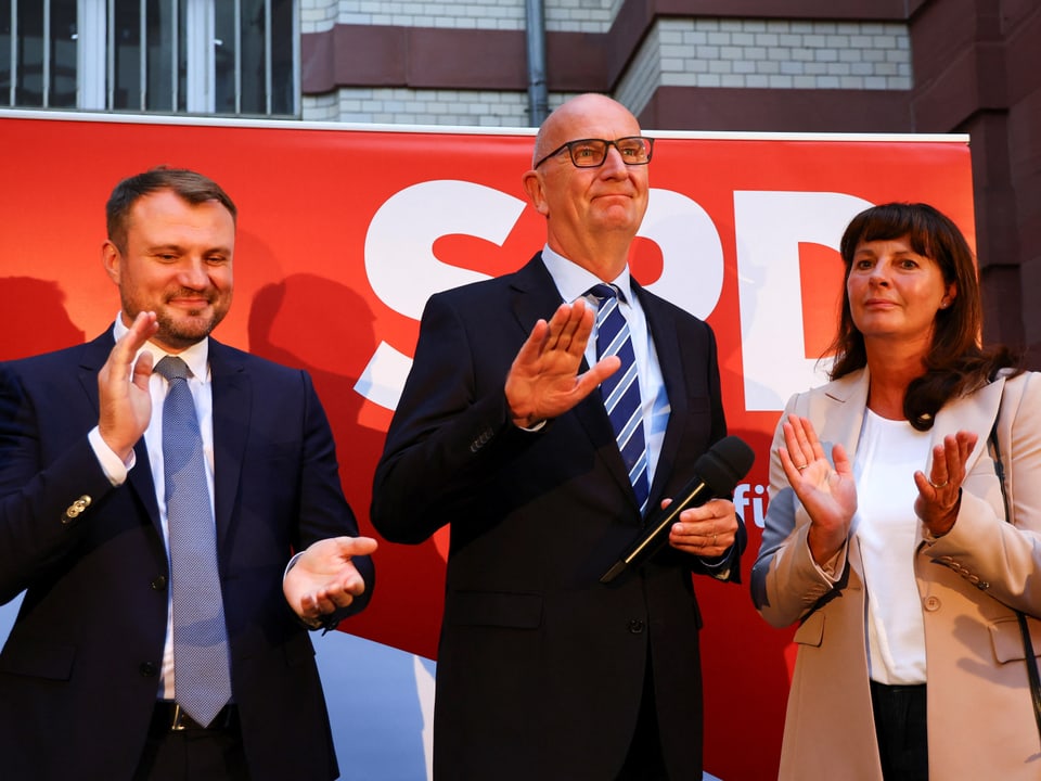 Drei Personen bei einer Veranstaltung vor einem roten SPD-Banner.