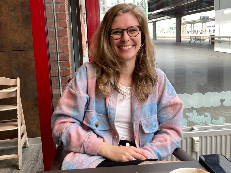 Lächelnde Frau mit Brille in einem Café sitzt vor einem Fenster.