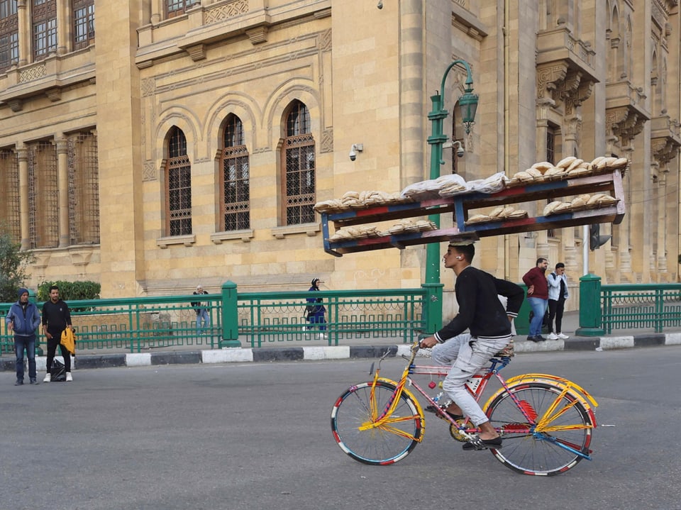 Mann auf Fahrrad mit Broten auf Kopfträger vor Gebäude.