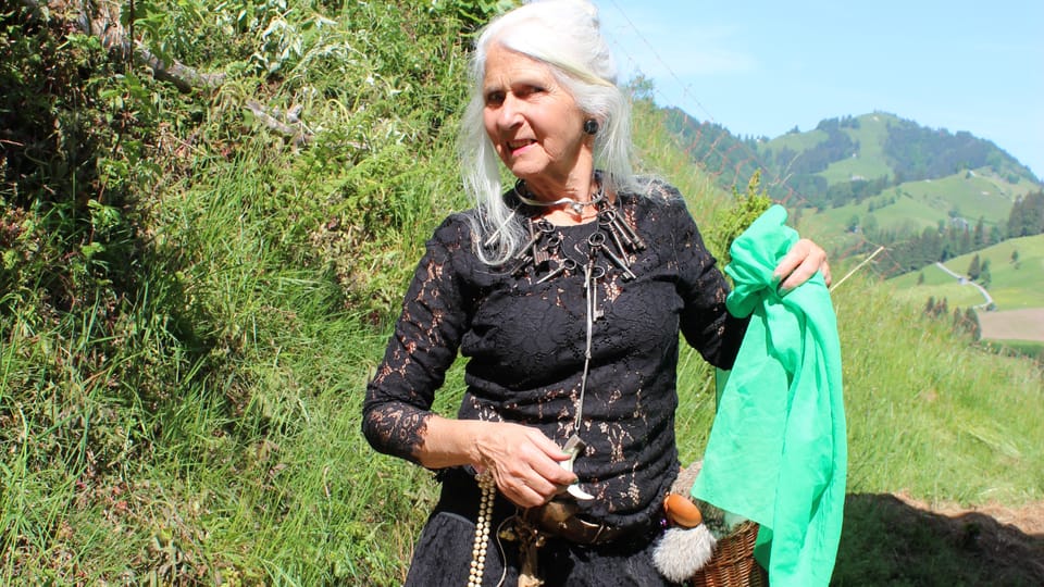 Frau mit grauen Haaren schwarzem Kleid und grünem Tuch in der Hand wandelt durch die Berglandschaft. 