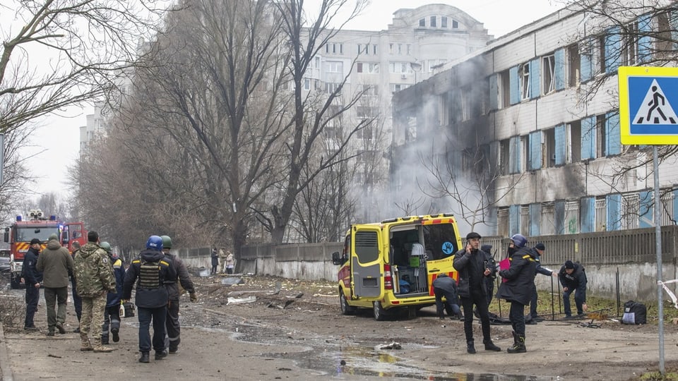 Bombardiertes Gebäude, davor Menschen und ein Krankenwagen.