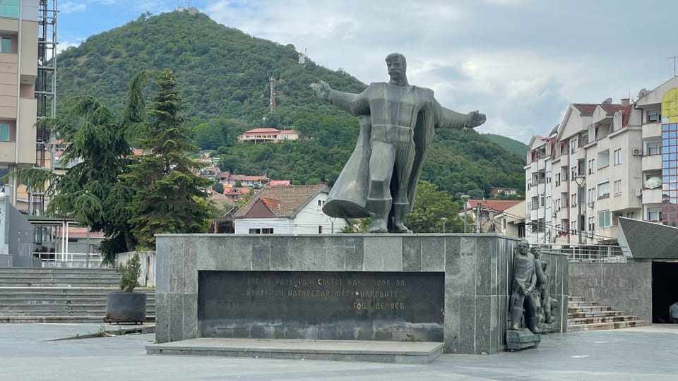 Statue eines Mannes mit ausgebreiteten Armen vor einem Hügel mit Bäumen.
