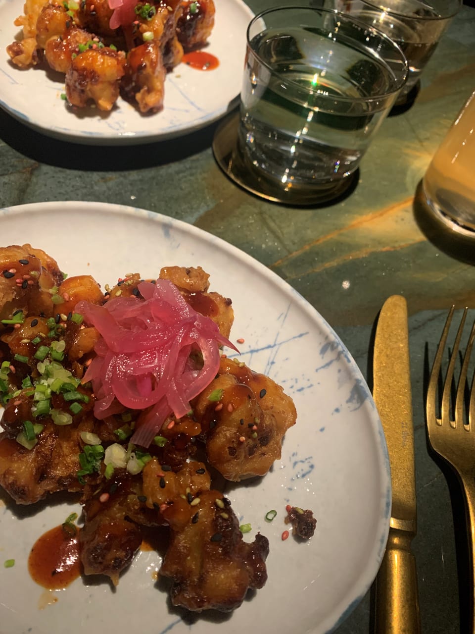 Teller mit frittierten Speisen, Zwiebeln und Sesam, daneben Besteck und Getränk.