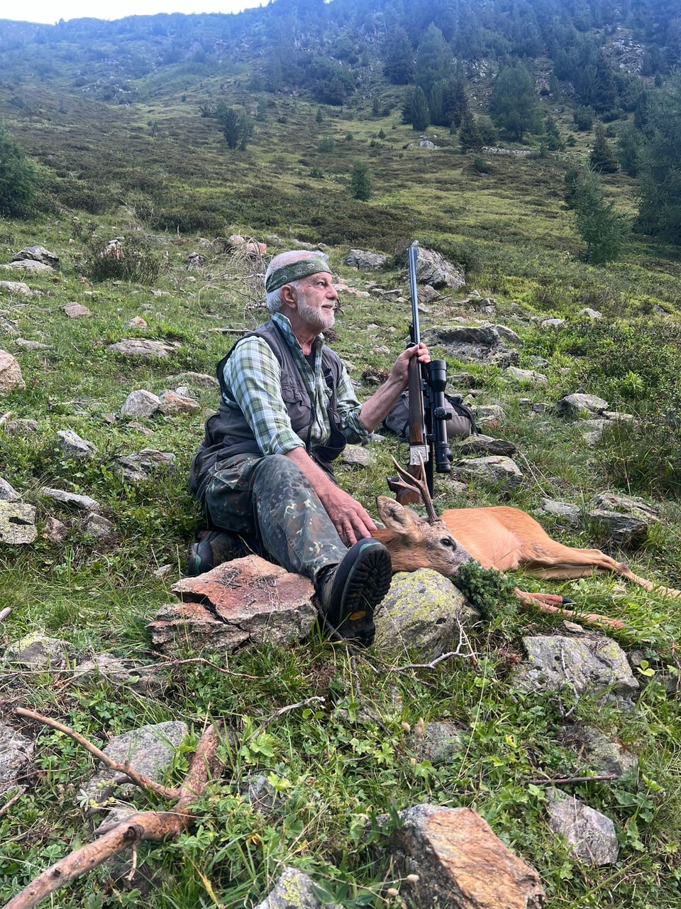 Jäger mit Gewehr und erlegtem Reh in bergiger Landschaft.