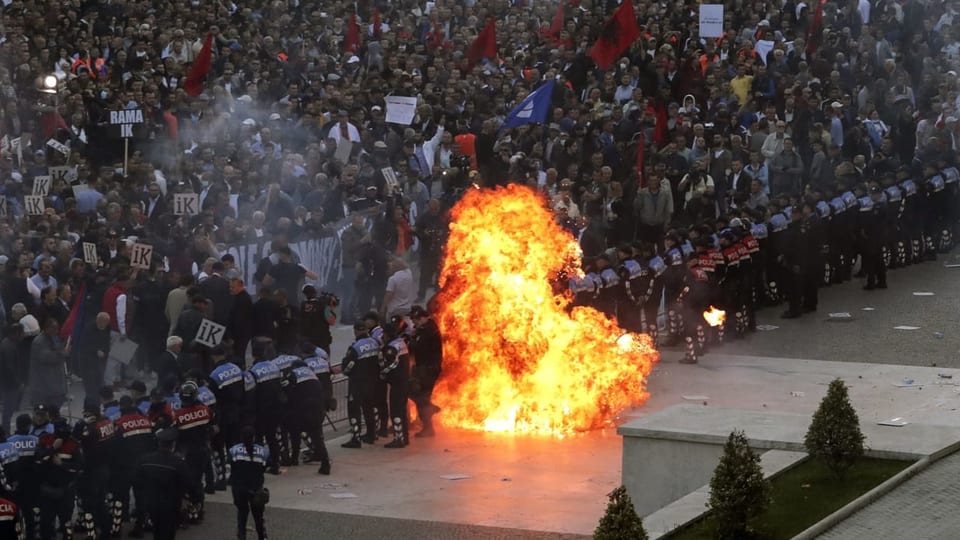 Blick auf zahlreiche Demonstranten. Davor eine Polizeikette und ein Molotow-Cocktail der explodiert und eine Feuerwolke erzeugt. 