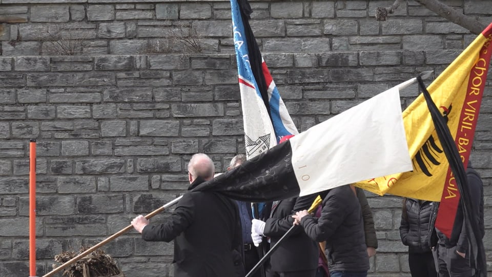 Menschen halten bunte Fahnen vor einer Steinmauer.