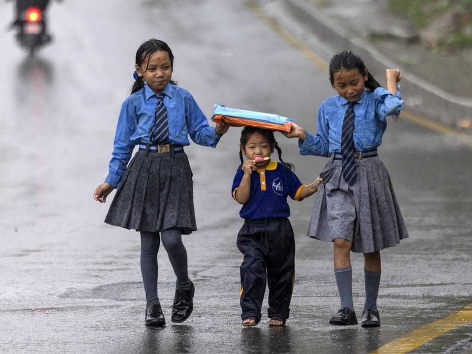 Drei Schulkinder laufen bei Regen auf einer Strasse, zwei halten ein Buch über das jüngere Kind.