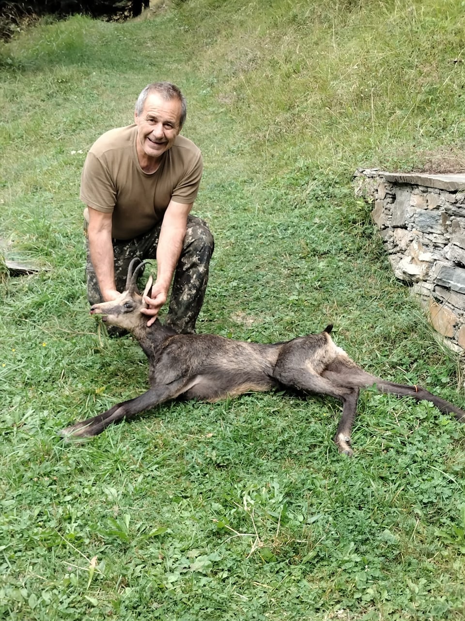 Mann posiert mit erlegter Gämse auf einer Wiese.