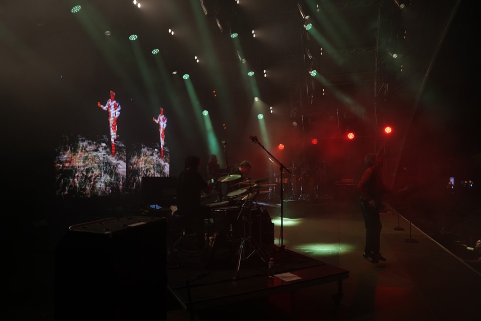 Band spielt auf Bühne mit Lichtern und Bildschirm im Hintergrund.