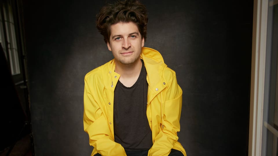 junger Mann mit dunbeklbonden Wuschelhaaren in knallgelber Jacke und schwarzem T-Shirt, Blick in Kamera.