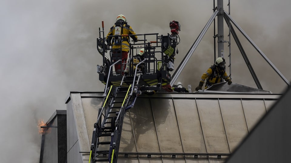 Feuerwehrleute auf dem Dach eines Bürogebäudes; dahinter ist Rauch zu erkennen