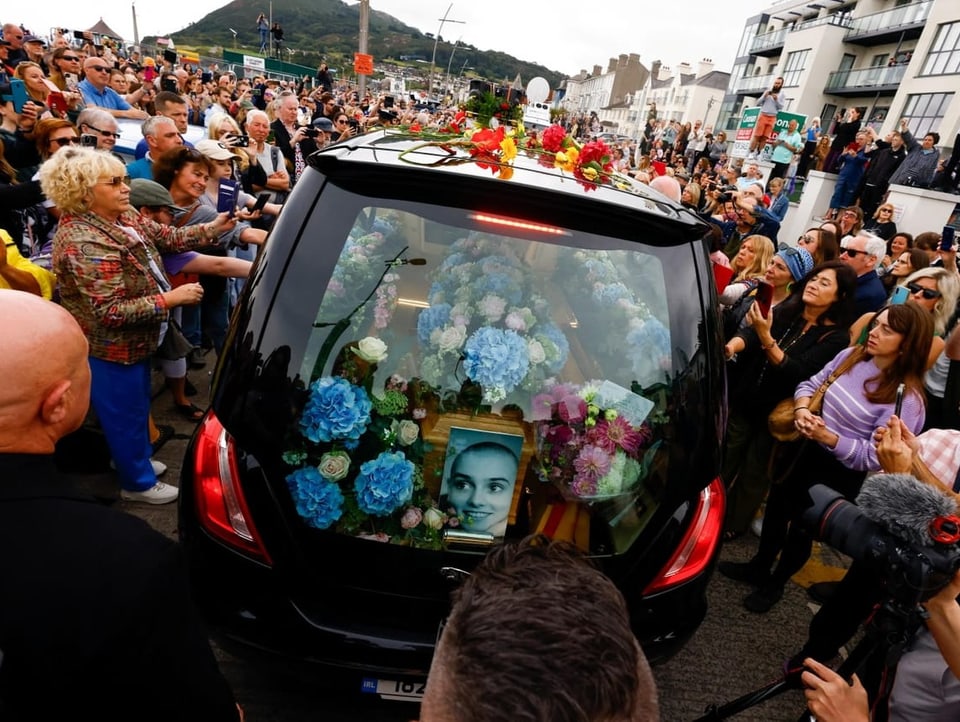 Blick von hinten auf den Leichenwagen mit Blumen und einem Bild der Sänderin; Dutzende stehen rundherum