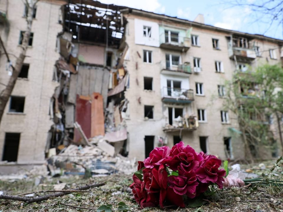 Blumen liegen auf dem Boden, im Hintergrund ein zerstörtes Gebäude.