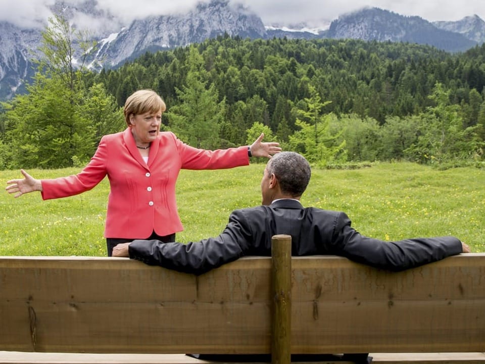 Angela Merkel hat die Arme weit ausgebreitet und steht vor Barack Obama, der auf einer Bank sitzt