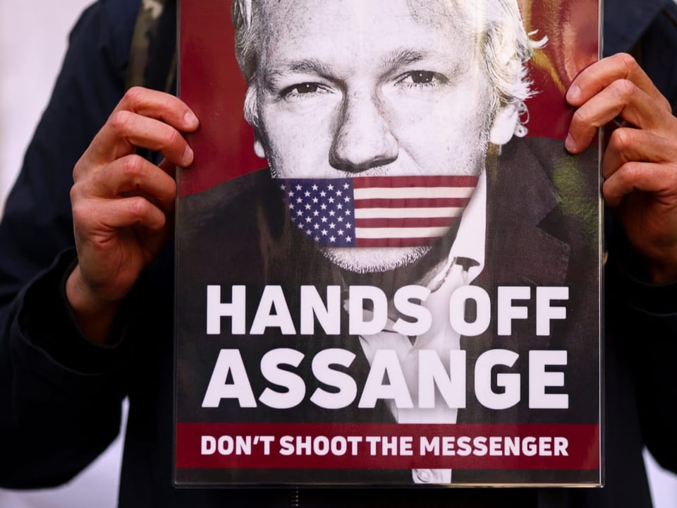 Eine Person hält ein Protestbanner in den Händen. Darauf das Gesicht von Assange und der Text: Hände weg von Assange.