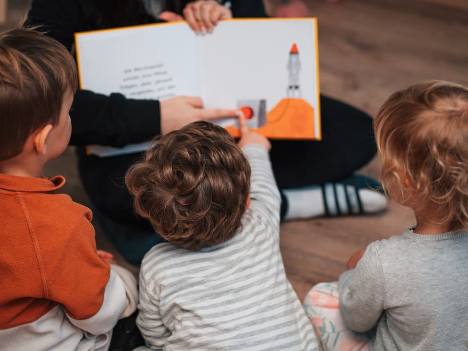 Drei Kinder sitzen vor einem aufgeschlagenen Bilderbuch.